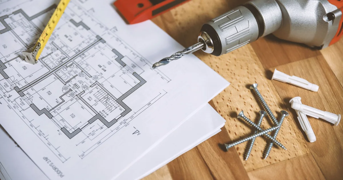 A printed house floor plan with a tape measure, a cordless drill, a spirit level, screws and wall plugs on a wooden table