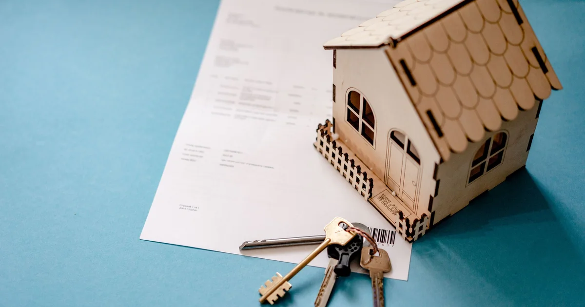 A small wooden house model with a set of keys and a signed contract on a blue table