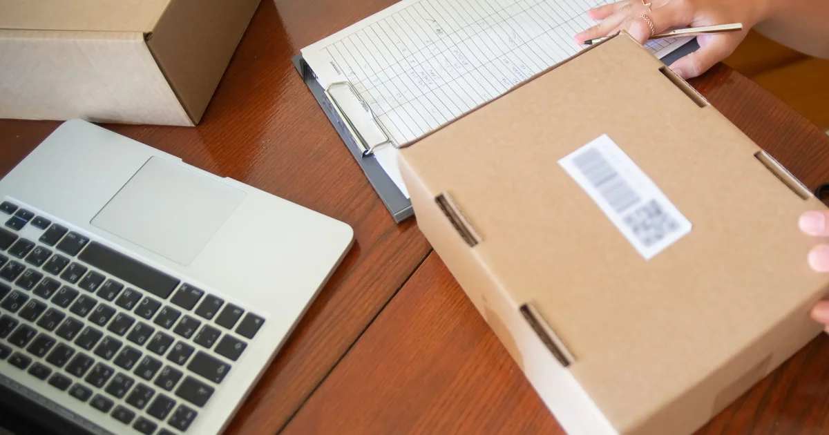 Online shop order being packed: a labelled parcel next to a laptop and an order list on a wooden table