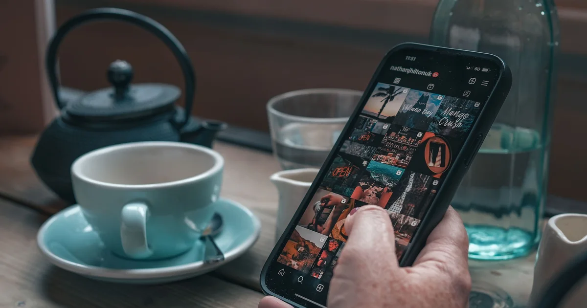 A hand holding a smartphone showing an Instagram grid, next to a teapot and a cup on a wooden café table