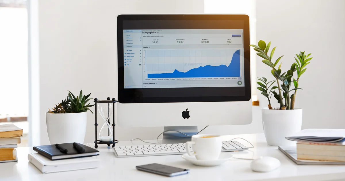 A desktop computer showing an analytics dashboard with a rising line chart, on a white desk with plants and a coffee cup