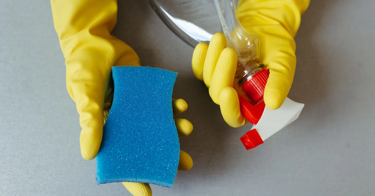 Hands in yellow rubber gloves holding a blue sponge and a spray bottle