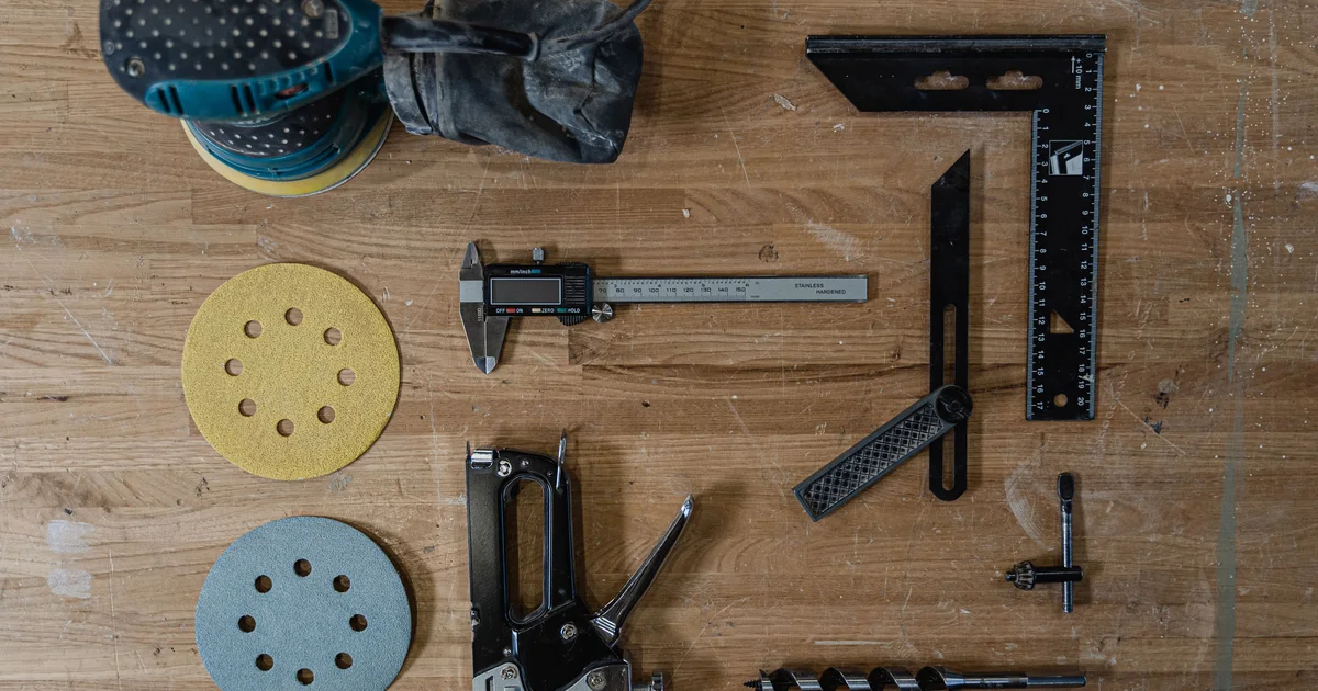 Woodworking tools laid out neatly on a wooden workbench: a sander, calipers, a square, a staple gun and sanding discs