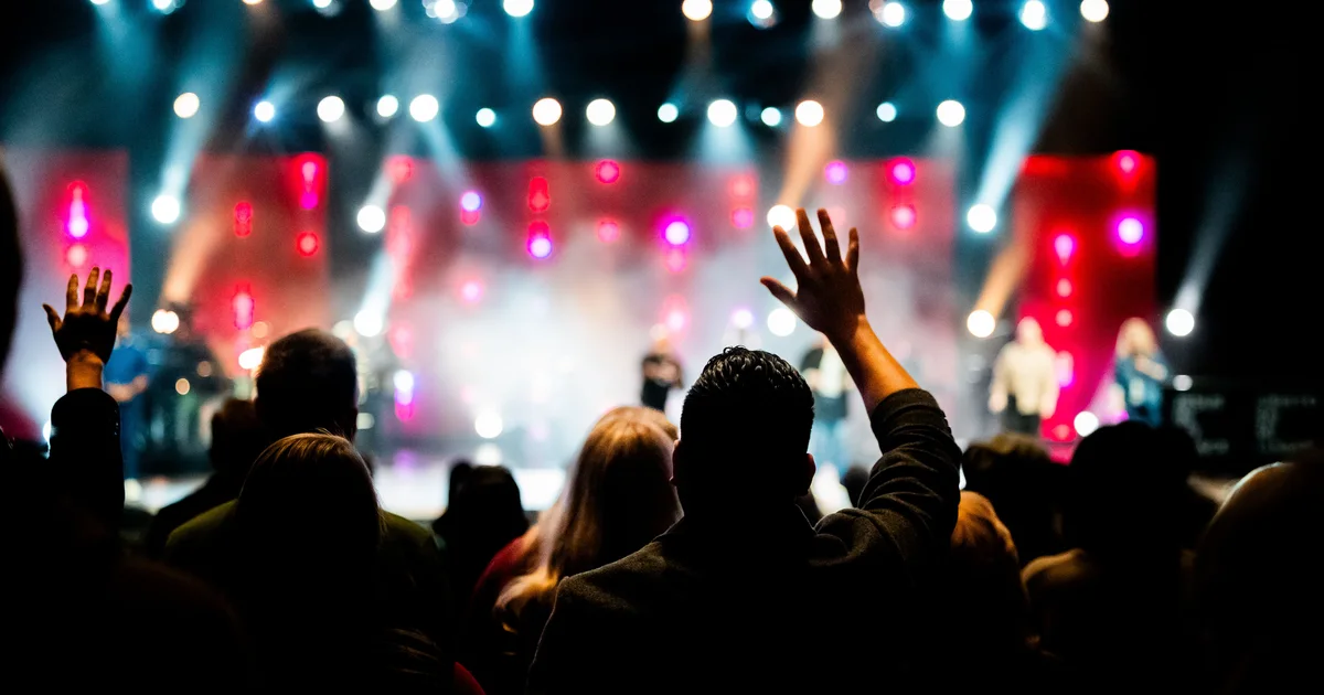 Silhouetted concert crowd with raised hands in front of a stage lit with bright white and red lights