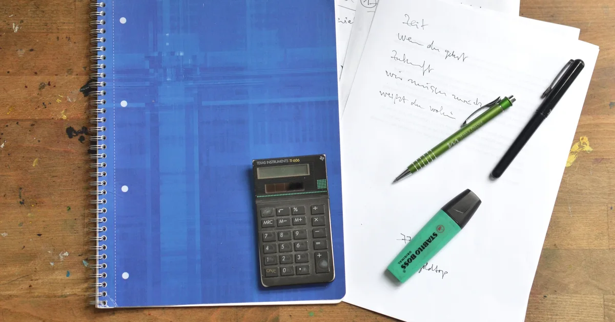 A spiral notebook, handwritten notes, a calculator, pens and a highlighter on a wooden desk