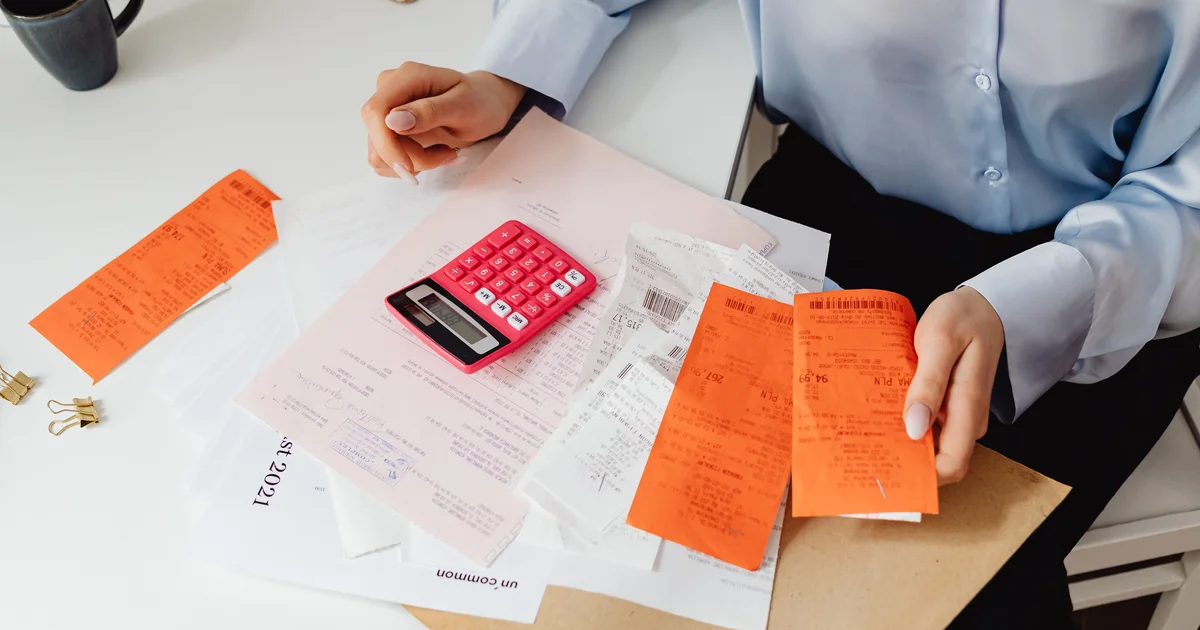 Hands sorting printed receipts and invoices next to a pink calculator on a white desk