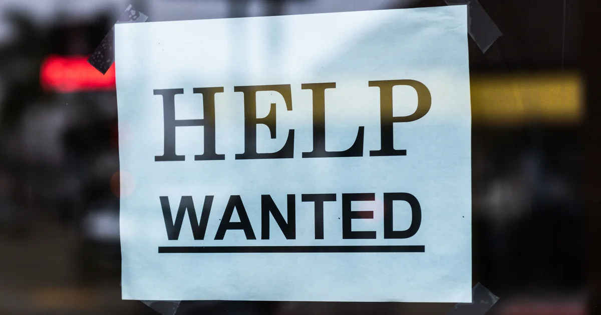 A printed Help Wanted sign taped to the glass window of a shop