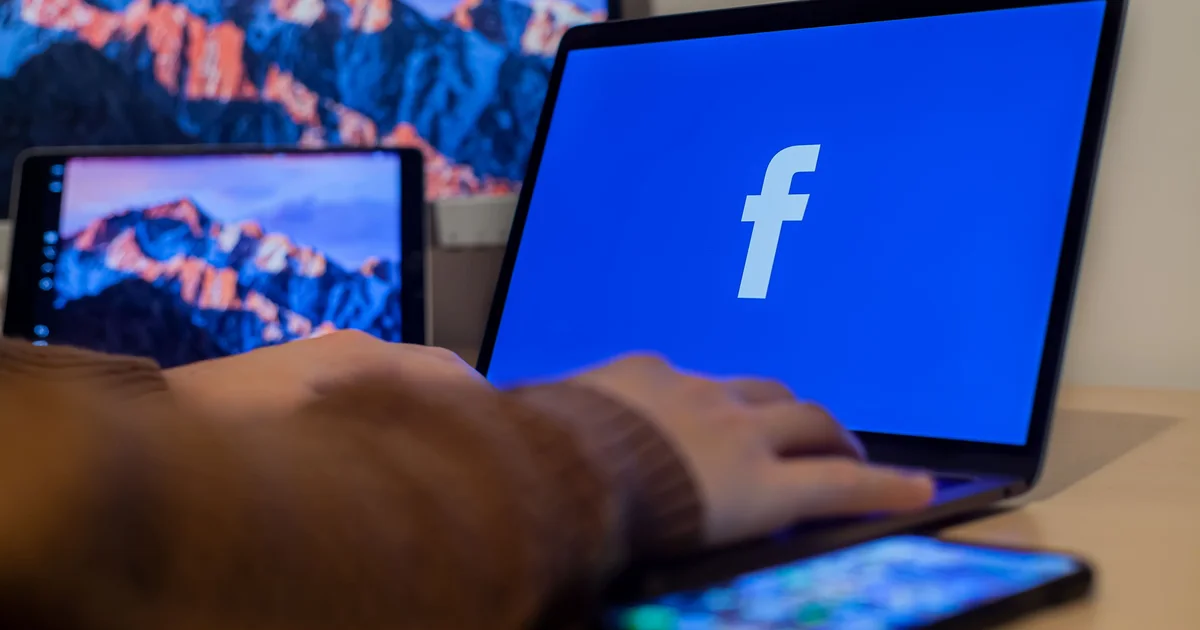 Hands typing on a laptop showing Facebook, with a smartphone beside it on the desk, the setting where Meta Advantage+ campaigns are built in Ads Manager
