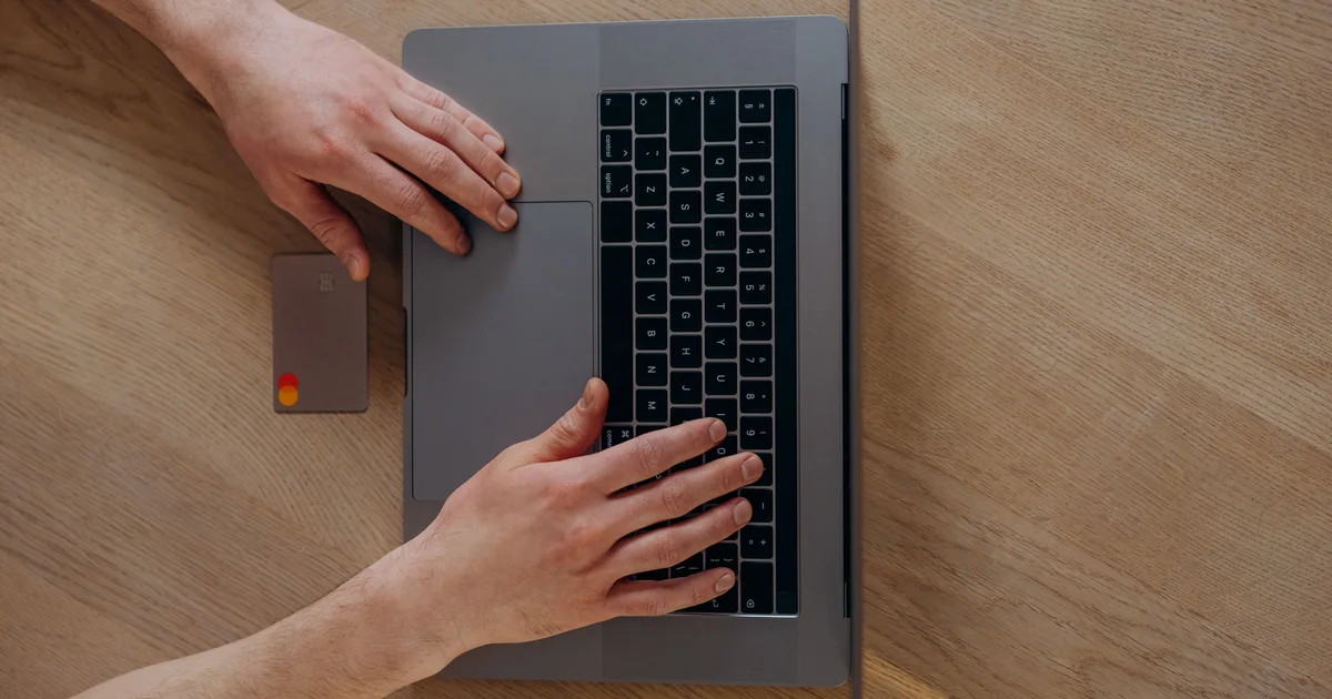Hands typing on a laptop on a wooden desk, with a bank card lying beside it