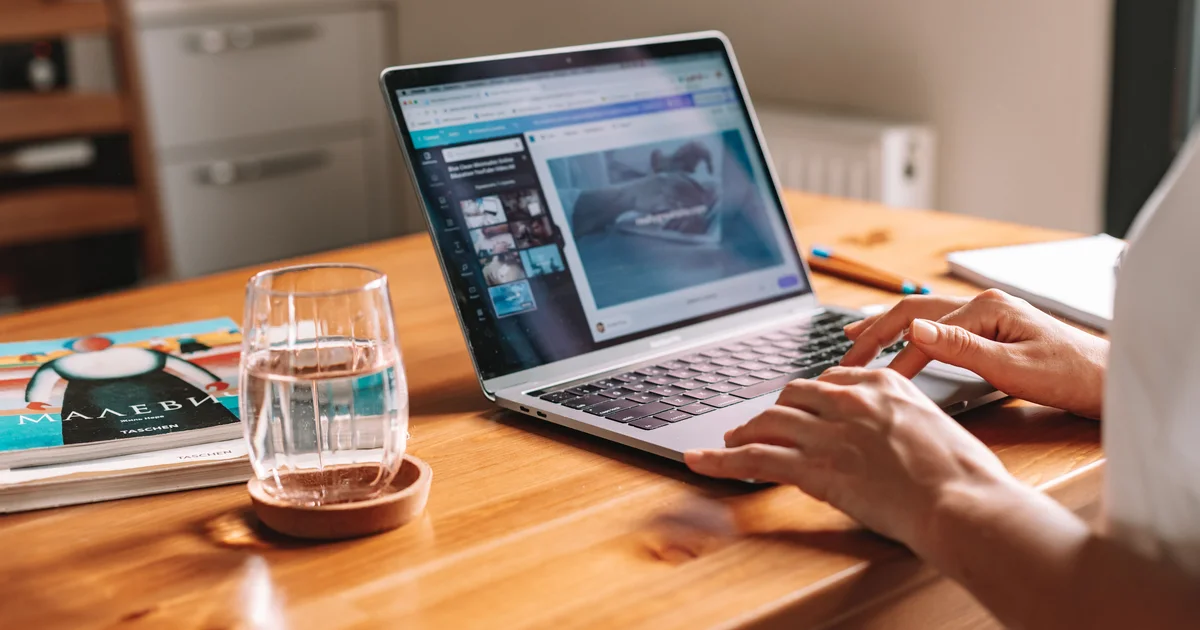 Hands typing on a laptop with a website being edited, on a wooden table next to a glass of water